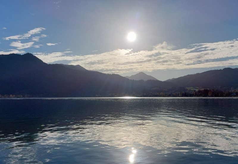 Das Bild zeigt einen Gebirgssee bei gleißendem Sonnenlicht. Im Hintergrund ist eine Bergkette zu sehen.