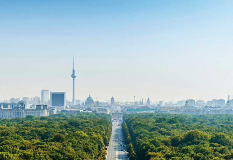 Das Bild zeigt die Straße des 17. Juni in Berlin aus der Luftperspektive. Rechts und links der Straße sieht man das Blätterdach der Bäume des Großen Tiergartens. Am Bildhorizont ist die Silouhette der Stadt zu erkennen, unter anderem mit Reichstagsgebäude, Fernsehturm, Dom und Brandenburger Tor.