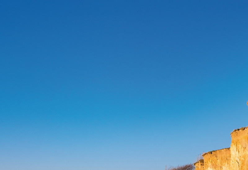 Das Bild zeigt die hellbraune Steilküste bei Ahrenshoop an der Ostsee. Am Fuß des Kliffs ist das Meer und die Gischt einer Welle zu sehen. An einem Küstenabschnitt befindet sich lockeres Material und Gestein, das sich von der Kliffoberkante an den Klifffuß verlagert hat.