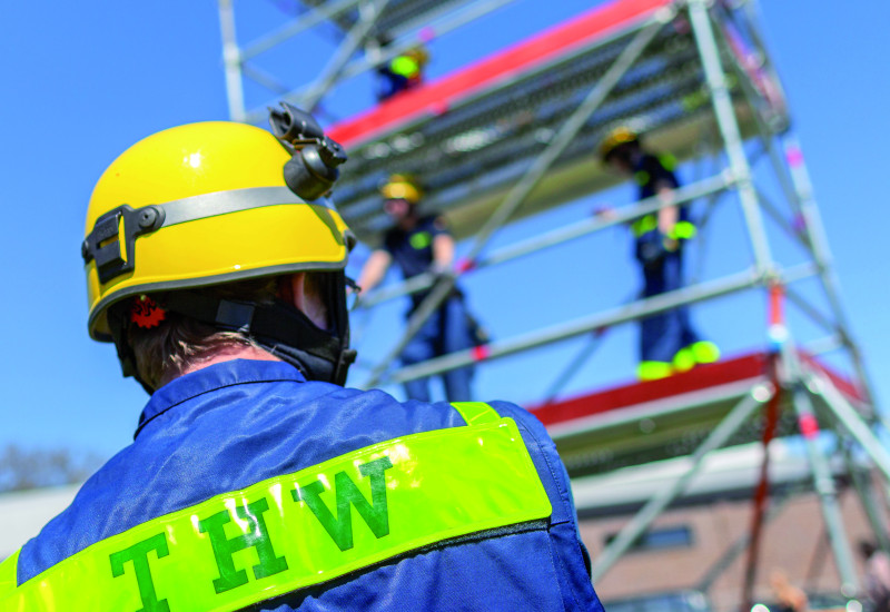 Das Bild zeigt einen Mitarbeiter des Technischen Hilfswerks in Uniform und mit Helm von hinten. Im Hintergrund ist ein Gerüst zu erkennen, auf dem weitere Mitarbeitende des THW stehen.