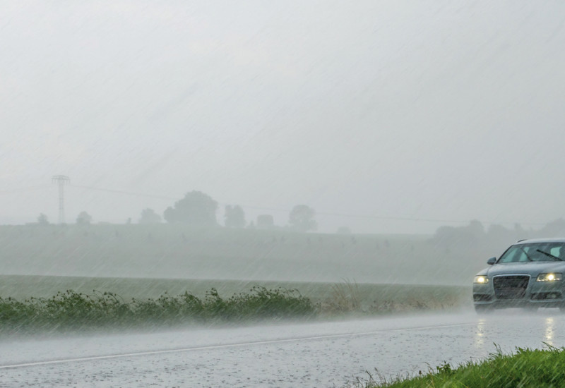Das Bild zeigt eine mit Wasser bedeckte Straße, auf der ein Auto fährt. Dieses hat Scheinwerfer und Scheibenwischer angeschaltet. Es regnet stark.