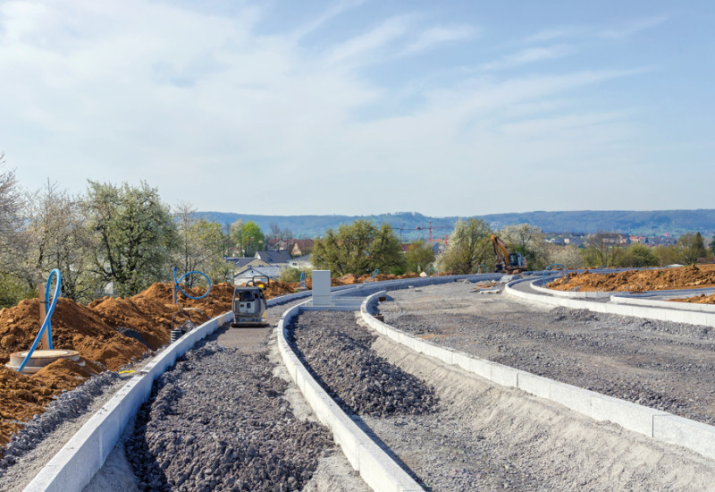 Das Bild zeigt einen Straßenneubau. Z sehen ist das teil-fertige Fundament für die Straße. Im Vordergrund ist es noch in Arbeit, im Hintergrund schon verdichtet. Links von der neuen Straße sind Erdhaufen zu sehen, dahinter eine Baumreihe. Im Hintergrund sind die Dächer eines Wohngebiets zu erkennen.