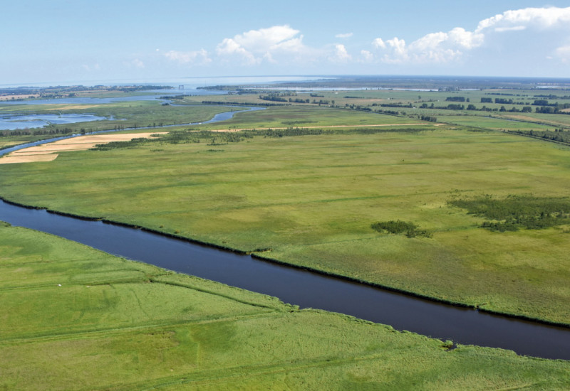 Das Bild zeigt einen Fluss, der durch eine flache Landschaft aus Wiesen und Landwirtschaftsflächen fließt. Im Hintergrund ist das Meer zu sehen.