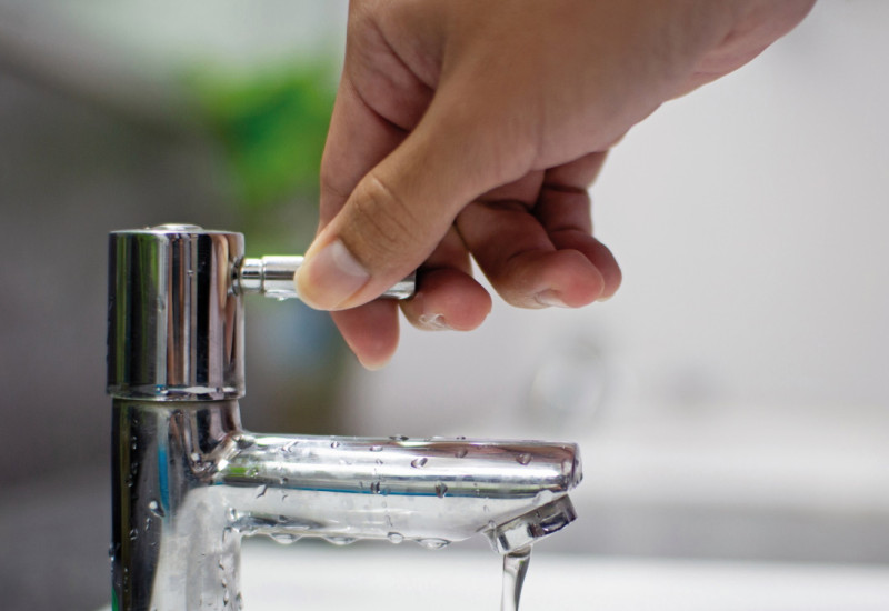 Das Bild zeigt eine Hand am Griff eines Wasserhahns, aus dem ein schwacher Wasserstrahl in ein Waschbecken fließt.