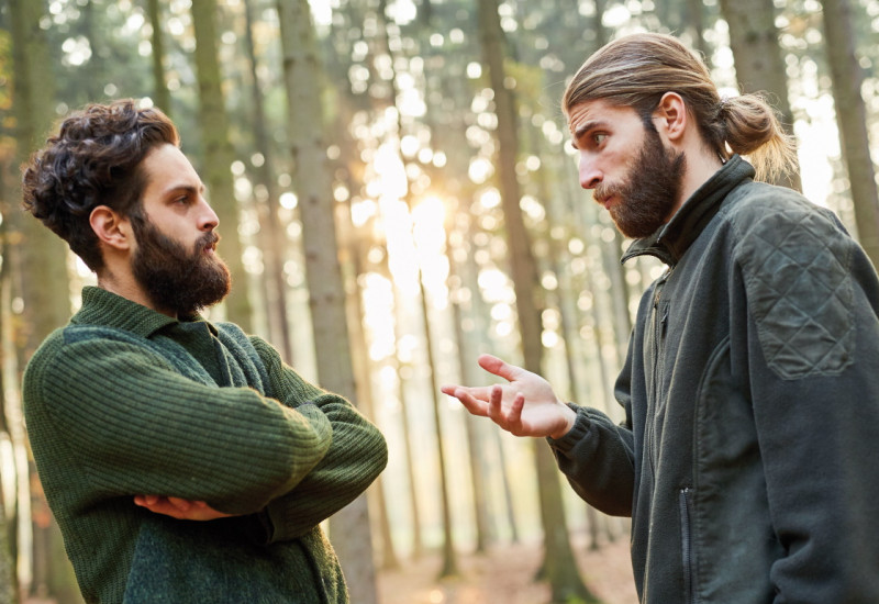 Das Bild zeigt zwei junge Förster, die in einem lichten Wald diskutieren. Der Mann links im Bild verschränkt die Arme, während der andere mit seiner Hand gestikuliert. Im Hintergrund dringen Strahlen der tiefstehenden Sonne in den Wald.