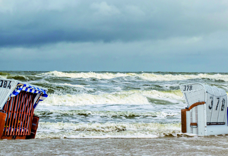 Das Bild zeigt einen Sandstrand an der Ostsee. Ein Strandkorb am Strand wird teilweise vom Wasser umspült. Im Hintergrund sind das Meer mit starkem Wellengang und dunkle Gewitterwolken zu sehen.