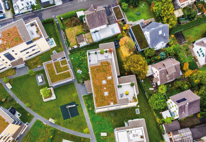 Das Bild zeigt eine Luftaufnahme eines Wohngebiets mit Ein- und Mehrfamilienhäusern. Zwischen den Gebäuden gibt es Rasenflächen und Bäume. Die Dächer der Mehrfamilienhäuser sind begrünt.