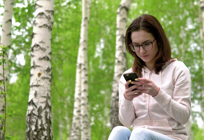 The picture shows a young women sitting in a birch wood. Her gaze is focused on a smartphone.