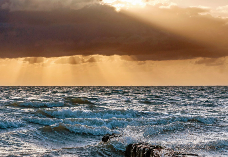 Das Bild zeigt den Ausschnitt eines unruhigen Meeres mit Wellen, die an aus dem Wasser herausragenden Pfählen brechen. Der Himmel ist orange gefärbt und von dunklen Wolken verhangen. Stellenweise treten Sonnenstrahlen zwischen den Wolken hervor.