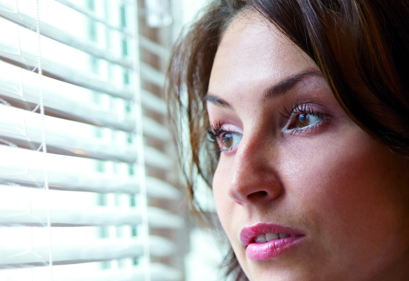 The picture shows a woman’s head and shoulders while she looks sceptically out of a window. The window is part-shaded by an indoor blind.