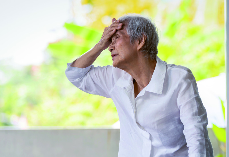 The picture shows an elderly woman wearing a white blouse. The lady is holding on to a rollator with one hand, while she is holding the back of her left hand to her forehead. In the background, a few fuzzy treetops or bushes are visible in bright light.