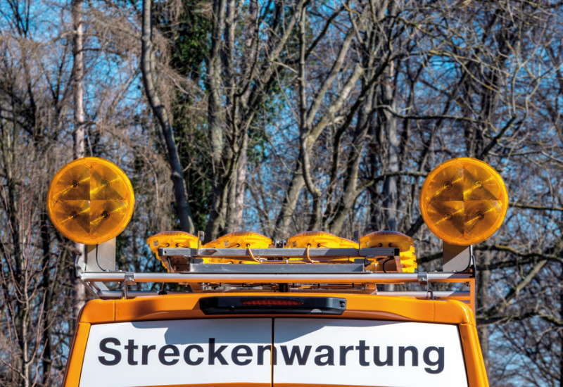 Das Bild zeigt das obere Teil des Hecks eines Fahrzeugs des Straßenbetriebsdienstes. Das Fahrzeug ist orange und seine Hecktüren sind mit dem Schriftzug "Streckenwartung" sowie rot-weiß gestreiften, reflektierenden Aufklebern beklebt. Auf dem Fahrzeug sind Warnlampen montiert.