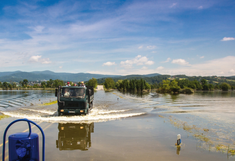 The picture shows an inundated landscape. In the centre of the picture a flooded road can be seen, with a lorry passing. There are people visible on the lorry’s load platform. In the picture’s foreground, part of a blue emergency vehicle is visible with blue warning lamps fitted all round the vehicle which is also driving along this flooded road.