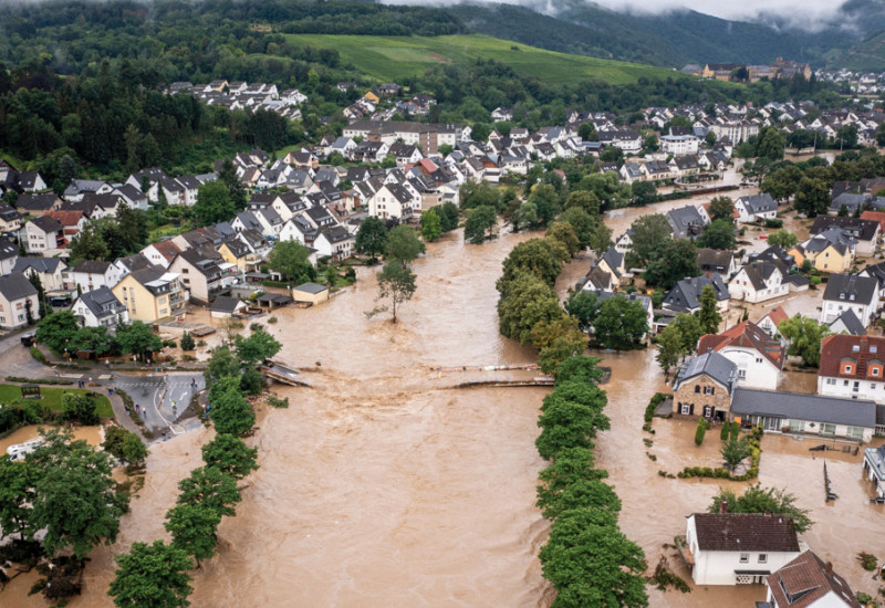 Das Bild zeigt eine Ortschaft mit Ein- und kleinen Mehrfamilienhäusern in einem Tal. Der durch die Ortschaft fließende Fluss ist über die Ufer getreten und überschwemmt die tiefer liegenden Teile der Ortschaft mit braunem Wasser. Der übliche Flusslauf ist durch Baumreihen zu erkennen. Zudem ist eine zerstörte Brücke, die den Fluss querte, sichtbar.