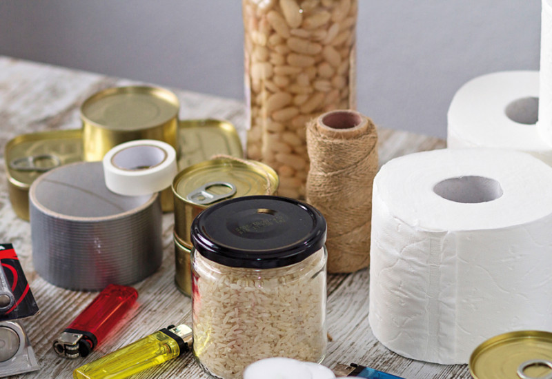 Das Bild zeigt eine Zusammenstellung von Notvorräten, darunter Lebensmittel wie Bohnen, Reis und Konserven, Batterien, Klebeband, Toilettenpapier, Teelichter und Feuerzeuge.