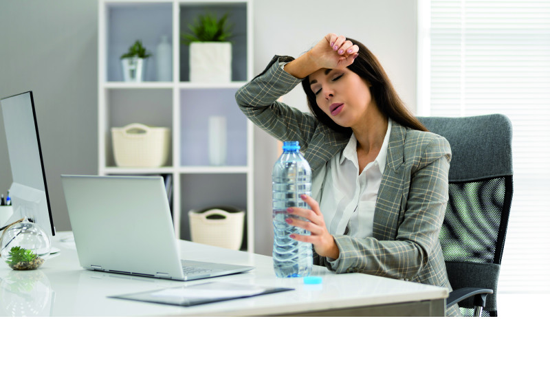 The picture shows a woman wearing a blouse and a blazer, sitting in an office in front of a laptop. In her left hand she is holding an open water bottle while wiping sweat off her brow with her right hand. The woman’s eyes are closed and her facial expression suggests exhaustion.