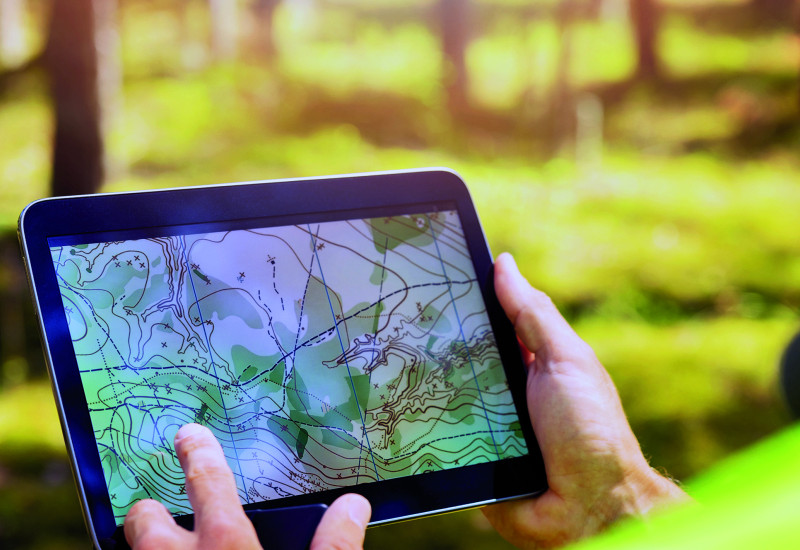 Das Bild zeigt Hände, die ein Tablet halten, auf essen Bildschirm eine Karte mit Höhenlinien und weiteren Markierungen zu sehen ist. Der verschwommene Bildhintergrund zeigt, dass die Person, die das Tablet hält, in einem Wald steht.