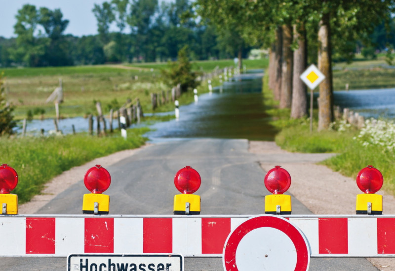 Das Bild zeigt eine durch eine Senke führende überflutete Straße. Im Vordergrund ist sie mit einer Straßensperre abgesperrt. An der Sperre hängt ein Schild mit der Aufschrift "Hochwasser". Rechts und links der Straße sind überflutete Wiesen zu erkennen.