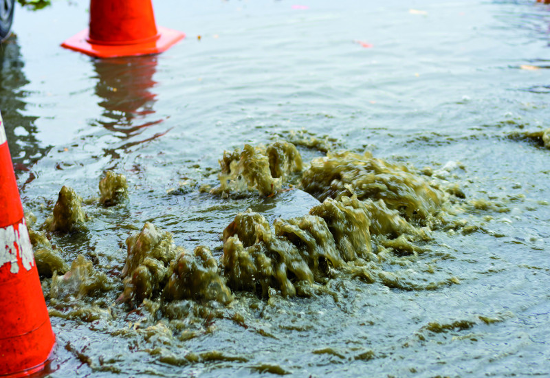 Das Bild zeigt einen Gully auf einer überfluteten Straße. Der Gully ist durch das daraus hervorsprudelnde Wasser erkennbar. Neben dem Gully stehen Warnkegel. Am äußersten Bildrand ist das Rad eines Autos zu erkennen.