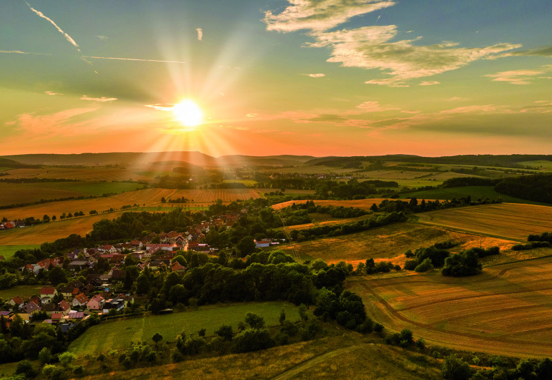 Das Bild zeigt eine leicht hüglige Landschaft, die ackerbaulich geprägt ist. In der Bildmitte ist ein Dorf in die Landschaft eingebettet. Am fast wolkenlosen, zum Teil schon orange gefärbten Himmel steht eine tiefstehende Sonne, die die Landschaft in warmes Licht hüllt.