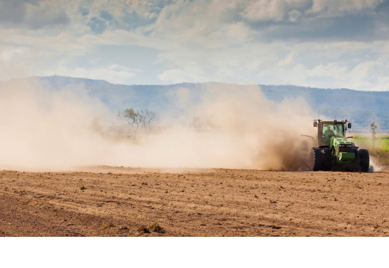 Das Bild zeigt eine trockene Ackerfläche, über die ein Traktor fährt. Aufgrund der Trockenheit wirbelt das Fahrzeug große Staubwolken auf. Im Hintergrund ist eine bewaldete Hügelkette zu erkennen.
