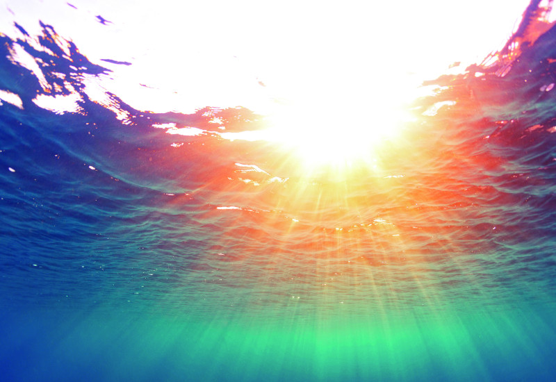 The picture shows a view of the surface of the sea from below. The sun’s rays are beaming through the water’s surface. The sea is coloured dark blue to turquoise-green.