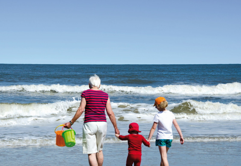 Das Bild zeigt eine ältere Frau, ein Kleinkind und ein Schulkind in Sommerkleidung von hinten. Die drei halten sich an den Händen und laufen am Strand gemeinsam auf die Wellen zu. Die Frau hält zwei Buddel-Eimer in der Hand.