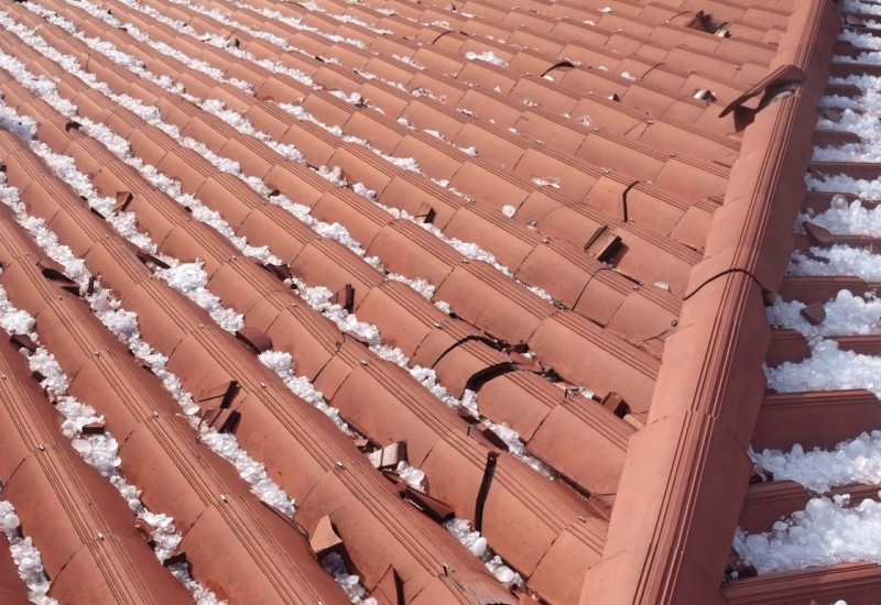 Das Bild zeigt ein mit roten Ziegeln gedecktes Dach von oben. Links und rechts des Dachfirsts liegen zahlreiche große Hagelkörner. Einige der Dachziegel sind zerbrochen.