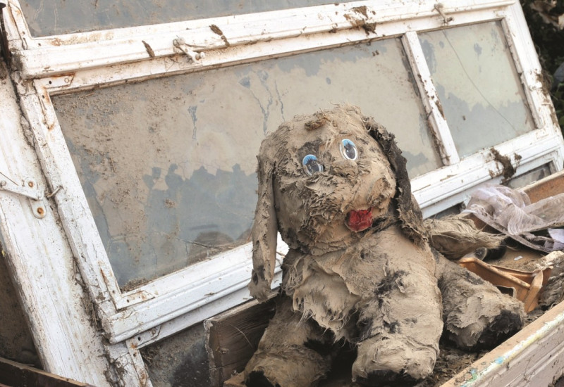 The picture shows piled up heavily soiled boards and windows. On top of the pile is a cuddly toy dog completely covered in mud.