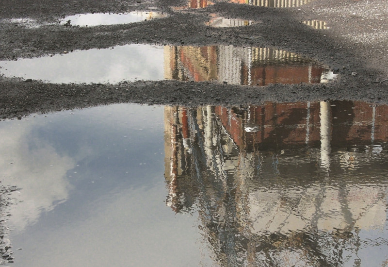 Das Bild zeigt zwei Pfützen auf einer Schotter-Kies-Fläche, in der sich ein Industriegebäude spiegelt.