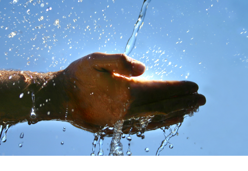 Zwei Hände werden in einen Wasserstrahl gehalten. Im Hintergrund scheint die Sonne