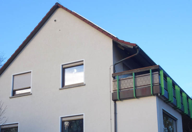 Das Gebäude von Südost mit dem Balkon über die gesamte Gebäudelänge, die Effizienz-Wärmepumpe steht unterhalb der linken Fenster