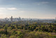 Looking over the top of a woodland, the picture shows the skyline of Frankfurt am Main. Skyscrapers and the tv tower rise high above the landscape. In the background, the Taunus mountain range is visible.