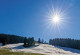 Das Bild zeigt einen Skilift, der auf eine überwiegend mit Nadelbäumen bewaldete Anhöhe führt. Eine dünne Schneedecke liegt auf der Anhöhe. Vor allem im Vordergrund sind aber weite Teile des Bodens ohne Schnee, sodass Rasen und nackter Boden zu erkennen sind. An einem wolkenlosen Himmel steht eine strahlende Sonne.