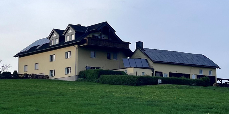 Außenansicht eines Gebäudes auf einer Wiese vor blauem Himmel, inkl Solarthermie und Photovoltaik auf dem Dach