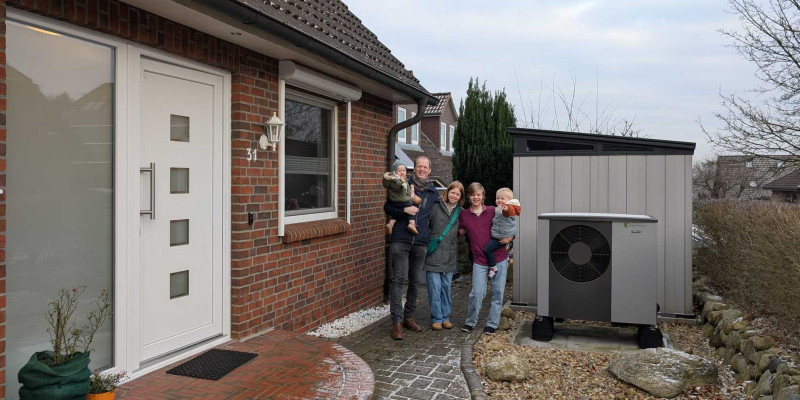 In der Bildmitte steht die fünfköpfige Familie, rechts neben ihr ist vor der Garage das Außengerät der Wärmepumpe zu sehen, links von ihr ein Teil des Hauses.