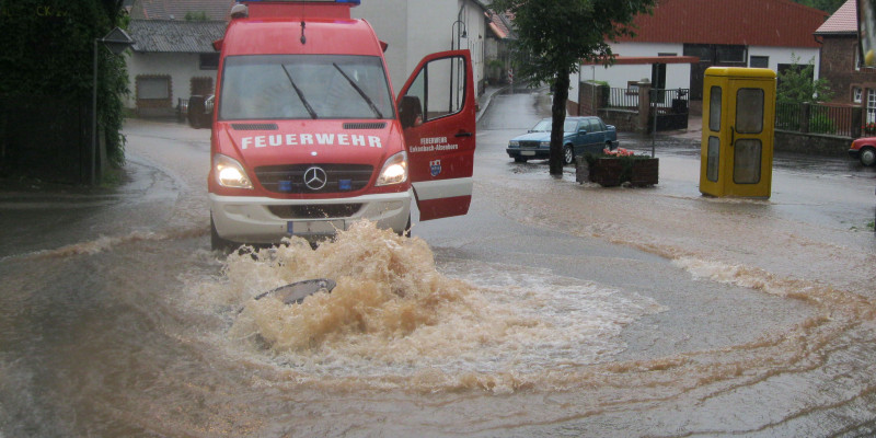 Fotografie einer überfluteten Straße. Der Gullydeckel wird von Wassermassen aus dem Boden gehoben. Das Wasser daraus fließt die Straße hinunter. Direkt vor dem Gullydeckel steht ein Einsatzwagen der Feuerwehr mit offener Tür.