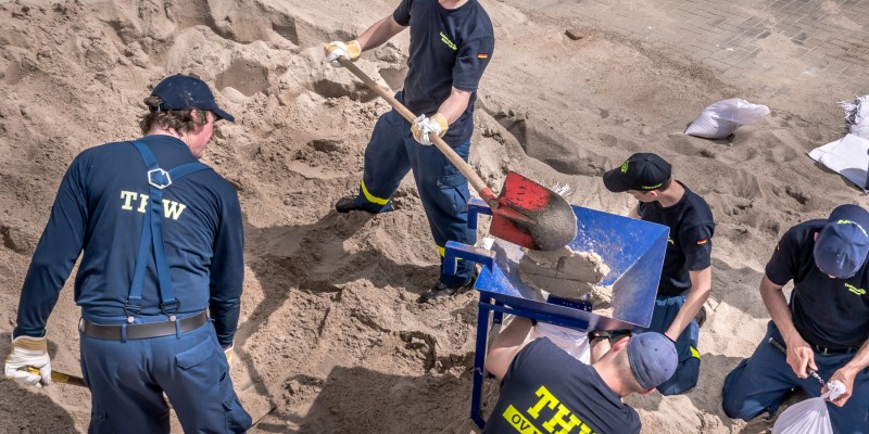 Ausbildungsteilnehmer_innen in Blau gekleidet und mit gelbem Schriftzug THW befüllen Säcke mit Sand mit Hilfe eines roten Spatens und einer blauen Schubkarre.