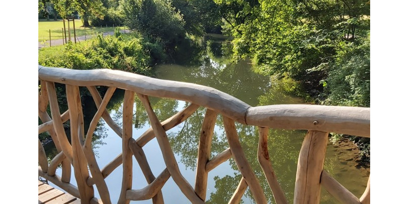 Fotografie von einer Holzbrücke auf einen darunterliegenden Bach neben einem Weg im Grünen.