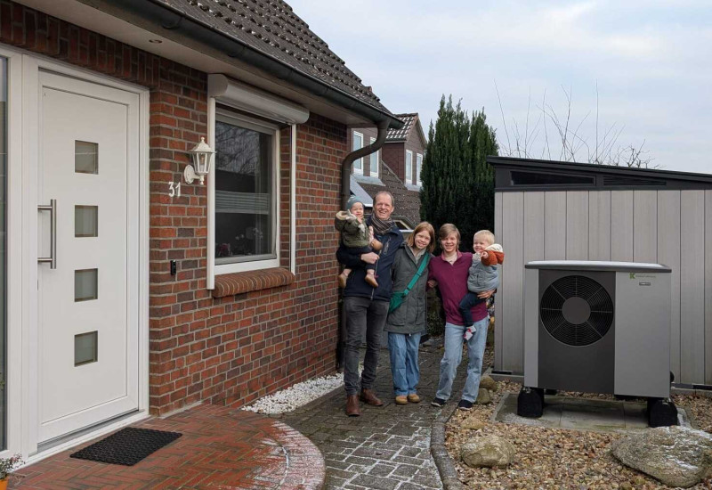 In der Bildmitte steht die fünfköpfige Familie, rechts neben ihr ist vor der Garage das Außengerät der Wärmepumpe zu sehen, links von ihr ein Teil des Hauses.