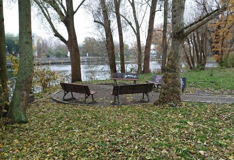 Parkbänke stehen kreisförmig zusammen unter unbelaubten Bäumen am Wasser. Im Hintergrund sind Bäume, Häuser und eine Brücke zu sehen.