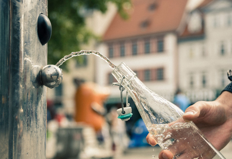 Eine Hand schöpft mithilfe einer wiederverwendbaren Glasflasche Wasser aus einem Trinkbrunnen in der Stadt.