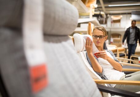 Eine junge Frau mit Brille sitzt in einem hellen Sessel in einem Möbelhaus und liest aufmerksam ein Produktetikett. Im Vordergrund ist ein grauer Sessel unscharf angeschnitten, während im Hintergrund Regale mit weiteren Möbeln und ein gehender Mann zu sehen sind.