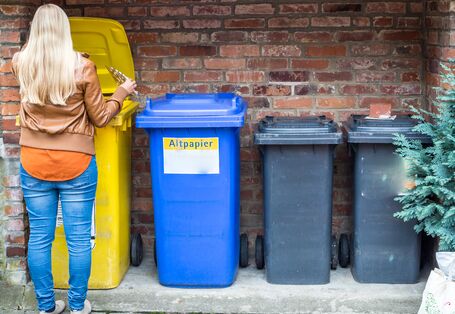 Eine Person mit blonden Haaren steht von hinten gesehen vor vier verschiedenen Mülltonnen (gelb, blau, zwei dunkelgraue) an einer Ziegelwand. Die Person öffnet gerade den Deckel der gelben Tonne, um eine leere Medikamenten-Blisterpackung einzuwerfen. Auf der blauen Tonne ist die Aufschrift „Altpapier“ zu erkennen.
