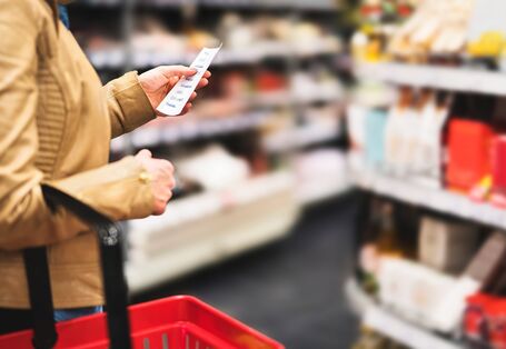 Eine Person in einer beigen Jacke und mit einem roten Einkaufskorb in der Hand hält in einem Supermarkt eine Einkaufsliste in die Höhe und blickt darauf.